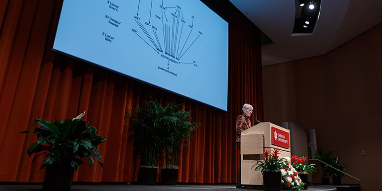 Kirsten Gronbjerg speaks at a podium, a presentation is lit up on a screen behind her